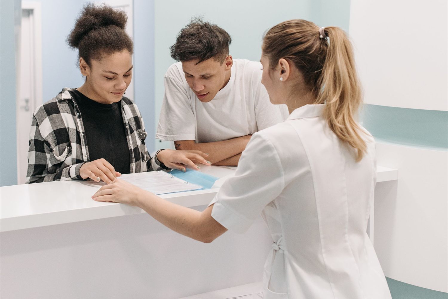 Eine MFA reicht zwei Patienten am Anmeldetresen einen Anamnesebogen auf Papier zum Ausfüllen.